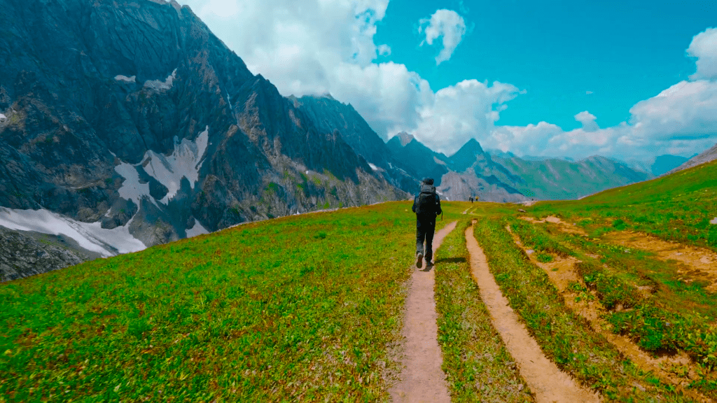 A breathtaking view of the serene Vishansar Lake, surrounded by lush green meadows and snow-capped peaks, captured during the Kashmir Great Lakes Trek. The clear blue sky reflects on the pristine waters, creating a perfect mirror image of the majestic mountains.